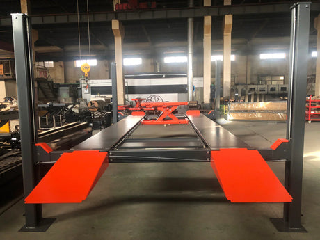 The Katool KT-4H110 4-post vehicle lift with gray posts and orange ramps is set up inside a warehouse, with a rolling jack and drip trays visible on the runways